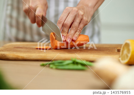 Person's hands preparing healthy food, slicing fresh carrot into thin pieces on a wooden cutting board in a kitchen 143576955