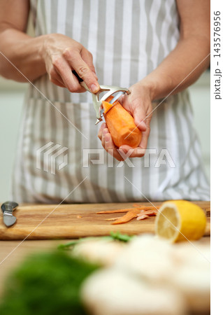 Hands peeling an orange carrot over a wooden cutting board with a vegetable peeler. Preparing fresh produce for cooking 143576956