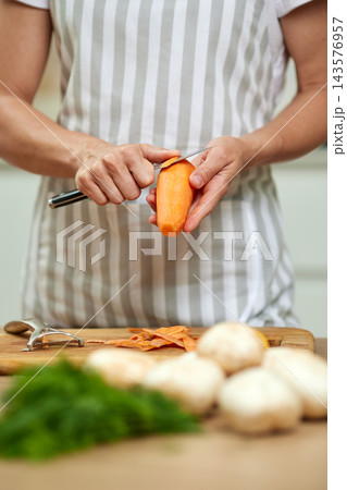 Woman's hands in a kitchen apron peeling fresh carrot with a peeler, preparing healthy vegetables on a wooden cutting board 143576957
