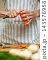 Person's hands peeling an organic carrot with a knife on a wooden chopping board, preparing healthy food in the kitchen 143576958