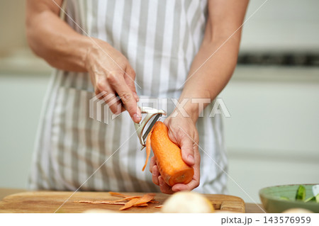 Person wearing apron peeling a raw carrot with vegetable peeler over a cutting board, preparing healthy food for cooking 143576959