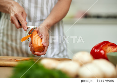 Hands of a woman chef peeling a fresh organic carrot over a wooden cutting board in a kitchen preparing a healthy meal 143576960