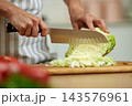 Person slicing green cabbage with knife on a wooden cutting board, preparing fresh vegetables in a kitchen 143576961
