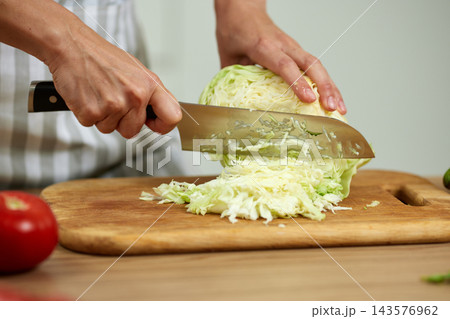 Hands cutting green cabbage with a sharp knife on a wooden cutting board, making a healthy vegetable salad 143576962