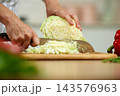 Person's hands using a knife to finely chop fresh green cabbage on a wooden board, preparing healthy food in a kitchen 143576963