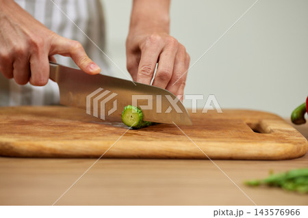 Chef hands preparing food, cutting fresh cucumber with a knife on a wooden chopping board in a modern kitchen 143576966