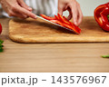 Hands slicing fresh red bell pepper into strips on a wooden cutting board, preparing healthy food in a kitchen environment 143576967