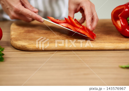 Hands slicing fresh red bell pepper into strips on a wooden cutting board, preparing healthy food in a kitchen environment Hands slicing fresh red bell pepper into strips on a wooden cutting board, preparing healthy food in a kitchen environment 143576967