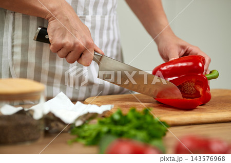 Hands in apron uniform cutting fresh bell pepper with a chef knife on a wooden board, preparing vegetable for cooking Hands in apron uniform cutting fresh bell pepper with a chef knife on a wooden board, preparing vegetable for cooking 143576968