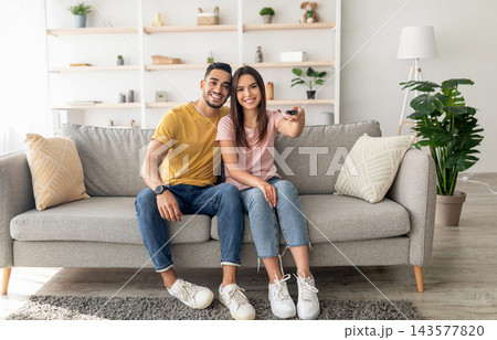 Joyful diverse couple watching TV program, sitting on couch at home, full length portrait. Cheery Arab man and his wife with remote control enjoying interesting movie in living room Joyful diverse couple watching TV program, sitting on couch at home, full length portrait. Cheery Arab man and his wife with remote control enjoying interesting movie in living room 143577820