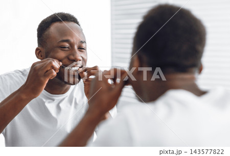 Toothy African Man Flossing And Cleaning Teeth Using Tooth Floss Standing Near Mirror In Modern Bathroom At Home In The Morning. Toothcare And Healthy Oral Hygiene Concept. Selective Focus 143577822