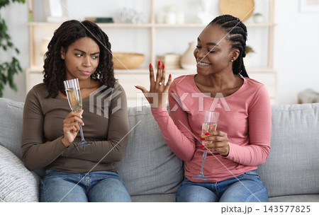 Happy woman showing her upset and jealous girlfriend engagement ring. Annoyed african american lady cannot share her friend happiness, drinking champagne together at home Happy woman showing her upset and jealous girlfriend engagement ring. Annoyed african american lady cannot share her friend happiness, drinking champagne together at home 143577825