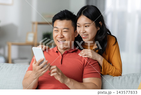 Cheerful chinese lovers having video chat with friends, positive asian couple making online party with family while staying together at home, using mobile phone, looking at gadget screen and smiling 143577833