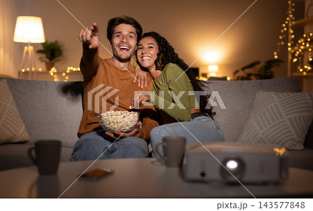 Joyful Couple Watching Film Online Using Home Cinema Projector Enjoying Evening Together On Weekend Sitting On Sofa In Living Room Indoors. Family Leisure, Entertainment And Fun Concept 143577848