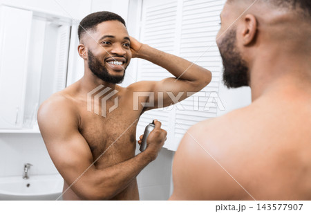 Smiling african american man with naked muscular torso using body spray, looking in mirror in bathroom. Man's daily care concept 143577907
