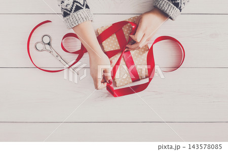 DIY hobby. Woman's hands wrapping christmas or other holiday handmade present in paper with red ribbon. Making bow at present box, decoration of gift. Scissors on white wooden table, top view. DIY hobby. Woman's hands wrapping christmas or other holiday handmade present in paper with red ribbon. Making bow at present box, decoration of gift. Scissors on white wooden table, top view. 143578085