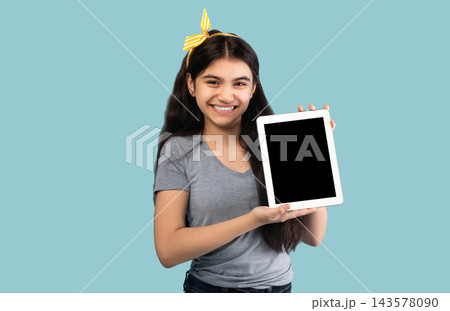 Cute Indian adolescent girl holding tablet pc with blank screen on blue studio background, space for app or website design. Smiling teenager showing touch pad with mockup for your ad Cute Indian adolescent girl holding tablet pc with blank screen on blue studio background, space for app or website design. Smiling teenager showing touch pad with mockup for your ad 143578090