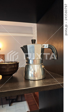 Stainless steel stovetop coffee maker beside a dark ceramic bowl on a kitchen shelf with warm light and contrasting neutral surfaces, still life 143580797