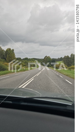 Empty winding road through lush countryside beneath dramatic gray storm clouds, viewed from inside a moving car with roadside markers and distant trees 143580798