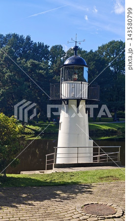 White lighthouse tower with dark domed lantern room and balcony beside a tree lined pond under blue sky in a landscaped park 143580799