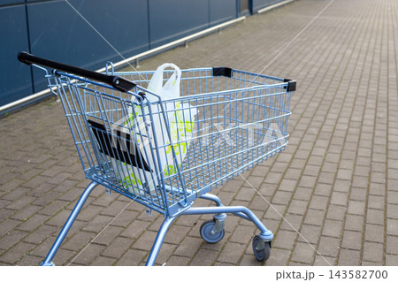 Light blue metal shopping cart carrying single plastic grocery bag stands on paved ground outside supermarket wall. Retail trade, consumerism, purchasing power, and grocery store service Light blue metal shopping cart carrying single plastic grocery bag stands on paved ground outside supermarket wall. Retail trade, consumerism, purchasing power, and grocery store service 143582700