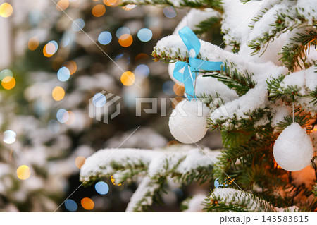 Snowy Christmas tree branches with white ball, blue ribbon on glowing fairy lights bokeh background Snowy Christmas tree branches with white ball, blue ribbon on glowing fairy lights bokeh background 143583815