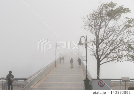 Famous Burgas pier bridge in thick fog over Black Sea. Iconic Burgas bridge shrouded in dense white fog, silhouettes of pedestrians walking along pier toward mist in Sea Garden park, Burgas, Bulgaria 143583892