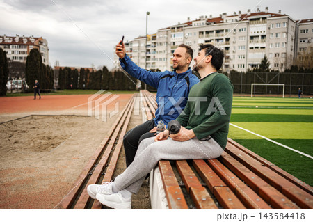 Male friends sitting on stadium benches taking selfie 143584418