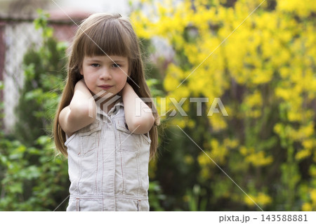 Portrait of a pretty little long-haired blond preschool girl in sleeveless white dress smiling shyly into camera against blurred outdoors background. Innocent happy childhood concept. 143588881