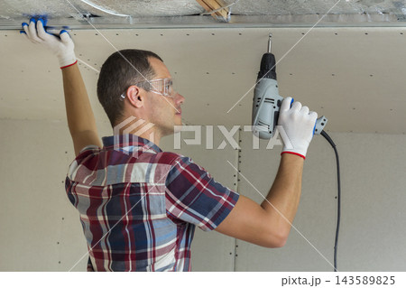 Young man in goggles fixing drywall suspended ceiling to metal frame using electrical screwdriver on ceiling insulated with shiny aluminum foil. Renovation, construction, do it yourself concept. Young man in goggles fixing drywall suspended ceiling to metal frame using electrical screwdriver on ceiling insulated with shiny aluminum foil. Renovation, construction, do it yourself concept. 143589825