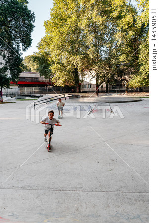 Children ride scooters at a skatepark during a sunny day in a city with trees and ramps 143590511