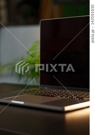 Laptop placed on a dark table next to a green plant in a room with soft lighting 143591030