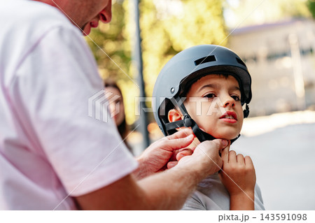 Child putting on helmet for safety while learning to ride a bike with adult support during the day Child putting on helmet for safety while learning to ride a bike with adult support during the day 143591098