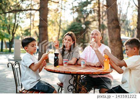 Family enjoys ice cream and drinks at a park on a sunny day in a casual setting with trees in the background 143591222