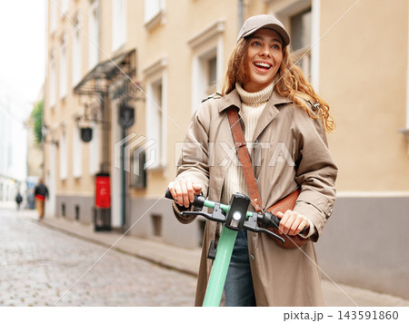 Woman rides a scooter on a city street with buildings and a smile on her face during daylight hours 143591860