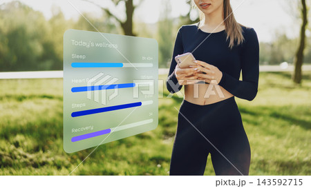 Woman checks wellness data on smartphone while standing in grassy area during daytime 143592715