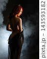 Red haired woman with crown and tattooed back draped in black sheer fabric, standing in smoke with dramatic backlighting 143593182