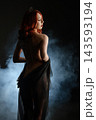Red haired woman with crown and tattoos draped in black sheer fabric against smoky dark background 143593194