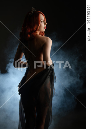 Red haired woman with crown and tattoos draped in black sheer fabric against smoky dark background 143593194