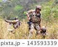 Three soldiers in full tactical gear advancing through tall grassy terrain with rifles ready, sunlight filtering through trees 143593339