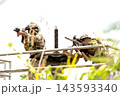 Two soldiers in camouflage gear aiming rifles from elevated metal platform amid bright foliage and overexposed sky 143593340