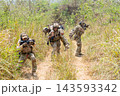 Soldier carrying wounded comrade through tall grassy trail with squad providing cover in sunlit jungle 143593342