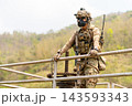 Soldier in full tactical gear leans on metal railing overlooking hilly jungle terrain with focused gaze 143593343