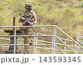 Soldier in full tactical gear stands alert on metal railing overlooking grassy hillside with rifle ready 143593345