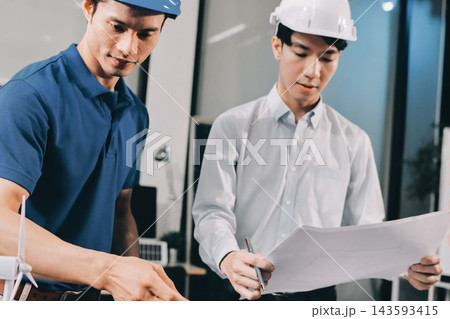 Two Asian engineers wearing hardhats and safety vests discussing solar panel installation and renewable energy project in the office. Two Asian engineers wearing hardhats and safety vests discussing solar panel installation and renewable energy project in the office. 143593415