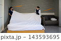 Two smiling professional women hotel housekeepers in uniform working together to spread clean white sheet over bed in  modern guest room 143593695