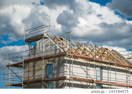 From the ground perspective, scaffolding supports the wooden trusses that form the roof structure of a building under construction, set against a dramatic cloudy sky. From the ground perspective, scaffolding supports the wooden trusses that form the roof structure of a building under construction, set against a dramatic cloudy sky. 143594075