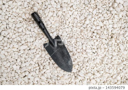 This gardening tool, a black hand trowel, is positioned on a bed of small white pebbles, ideal for various gardening tasks. 143594079