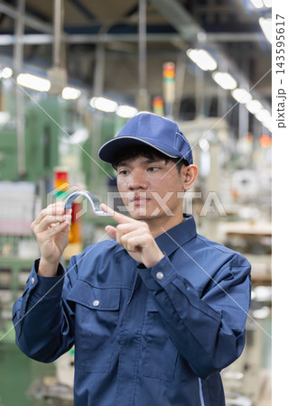 製造工場でプレス加工された金属部品を手に持ち、品質や仕上がりを目視確認する外国人男性労働者 143595617