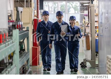 製造工場の通路を正面に向かって歩くベテラン男性技術者と若手男女作業員の3人チーム 143595731